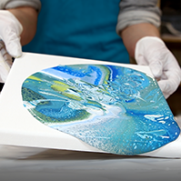 Geneigte Leinwand mit blauem und gruenem Acryl Pouring Farbverlauf in den Haenden gehalten