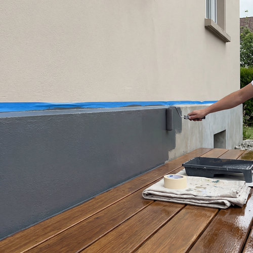Eine Person streicht den unteren Bereich einer Hausfassade mit grauer Farbe, die Kante ist mit blauem Abklebeband sauber abgeklebt, Farbwanne und Malerzubehör liegen bereit.