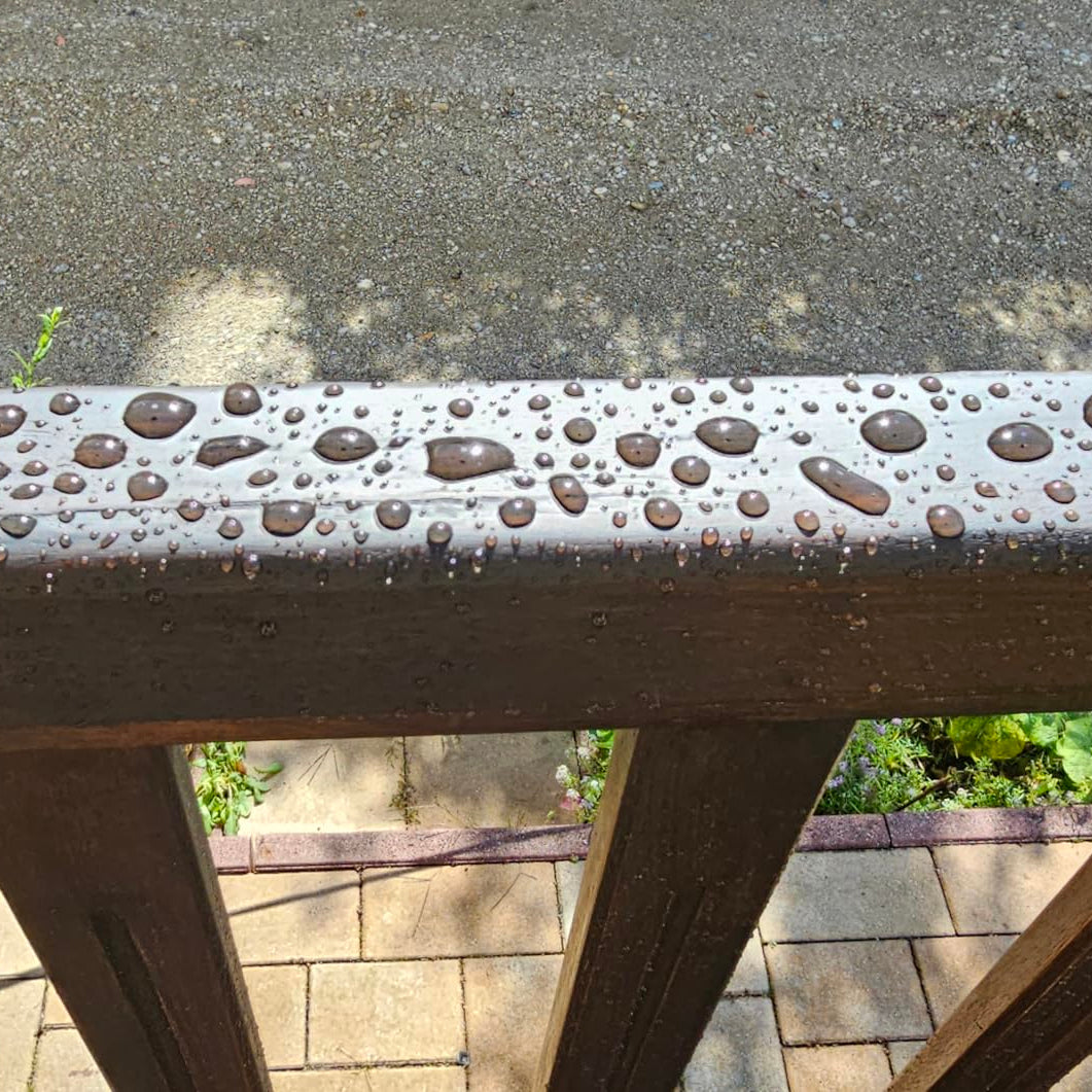 Nahaufnahme eines lasierten Holzgeländers mit abperlenden Wassertropfen als Zeichen wasserabweisender Wirkung.