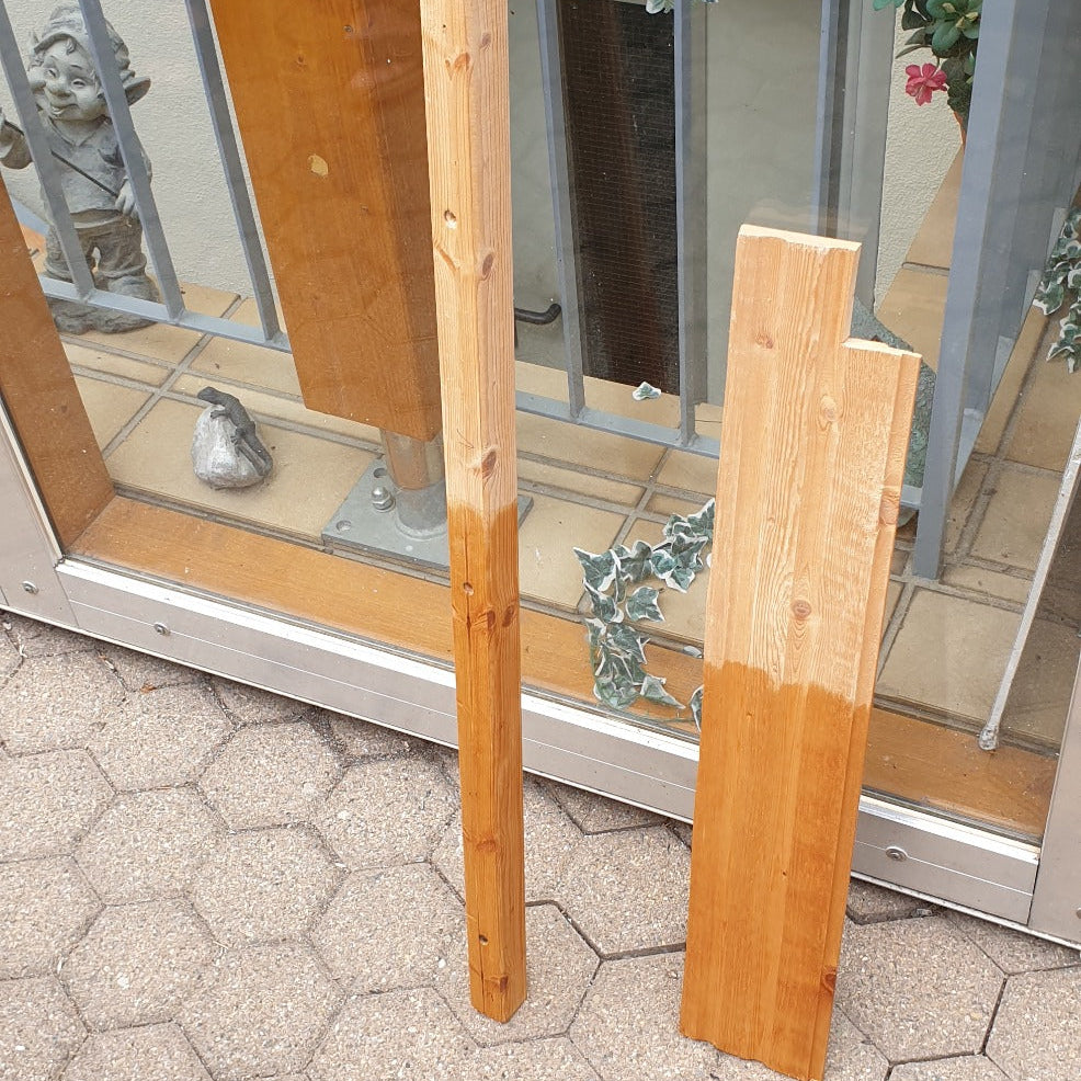 Zwei Holzpfosten im Außenbereich mit teilweiser Behandlung, die den Unterschied zwischen unbehandeltem und geöltem Holz zeigen.