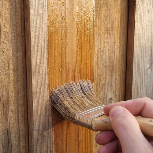 Nahaufnahme einer Hand, die mit einem Pinsel Holzschutzlasur auf senkrechte Holzbretter aufträgt, sichtbar sind frische Lasur und Wassertropfen auf der Oberfläche.
