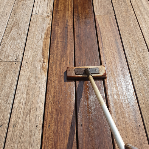 Holzdielen einer Terrasse beim Einölen, eine Bürste verteilt dunkles Holzöl auf den Brettern und zeigt den Farbunterschied zwischen behandelten und unbehandelten Flächen.