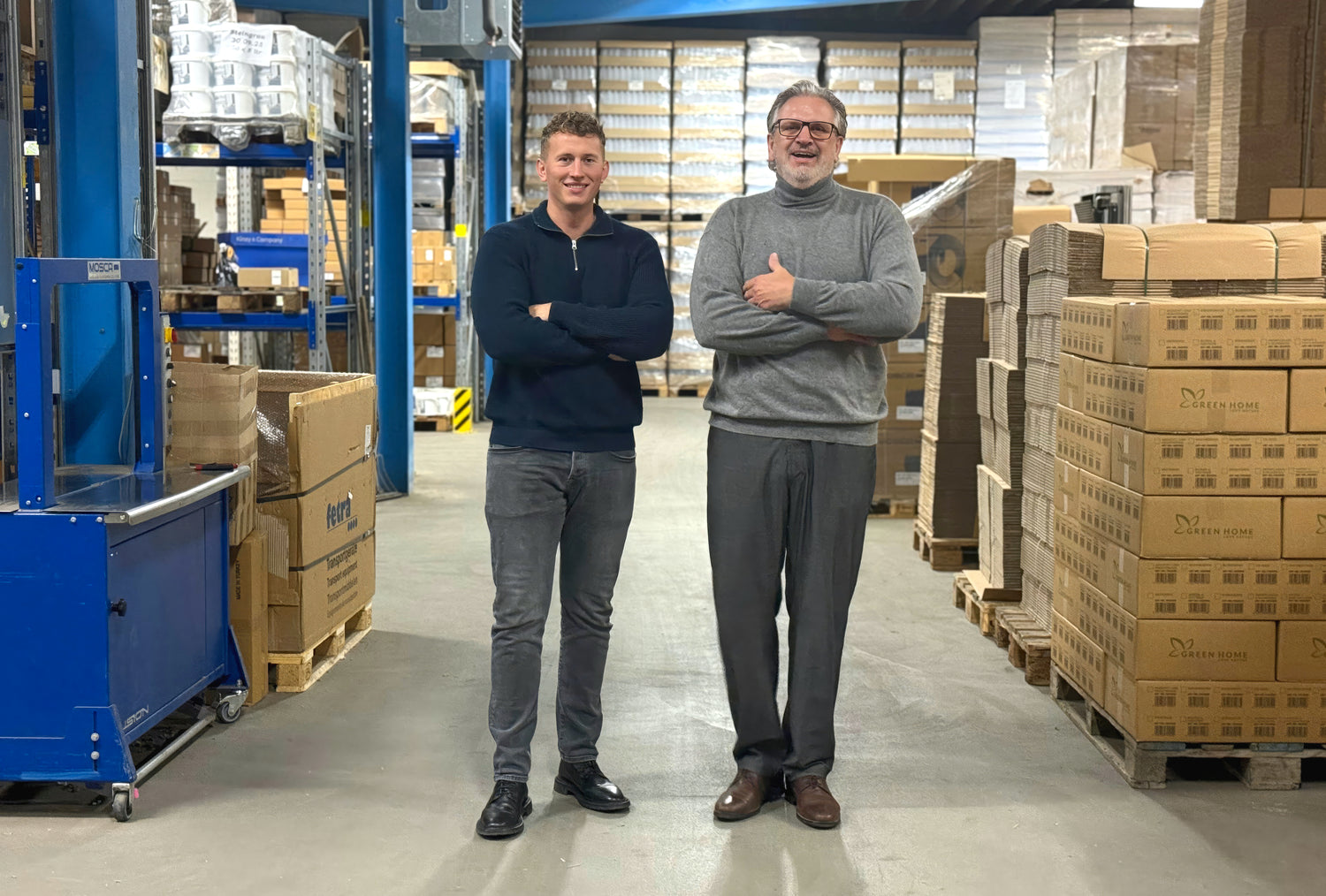 Zwei Männer stehen mit verschränkten Armen in einer Lagerhalle zwischen Paletten mit Kartons und hohen Regalen voller Waren.
