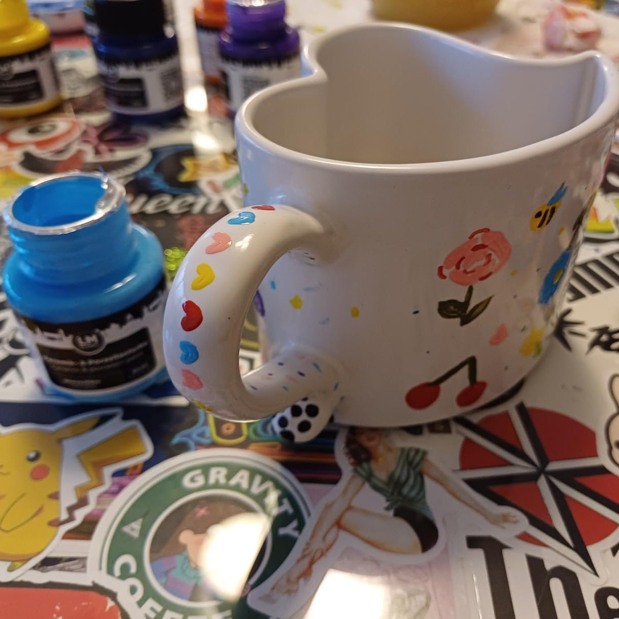 Mit LM Kreativ Keramik- und Porzellanfarbe gestaltete Herzhenkel-Tasse mit bunten Motiven auf Basteltisch