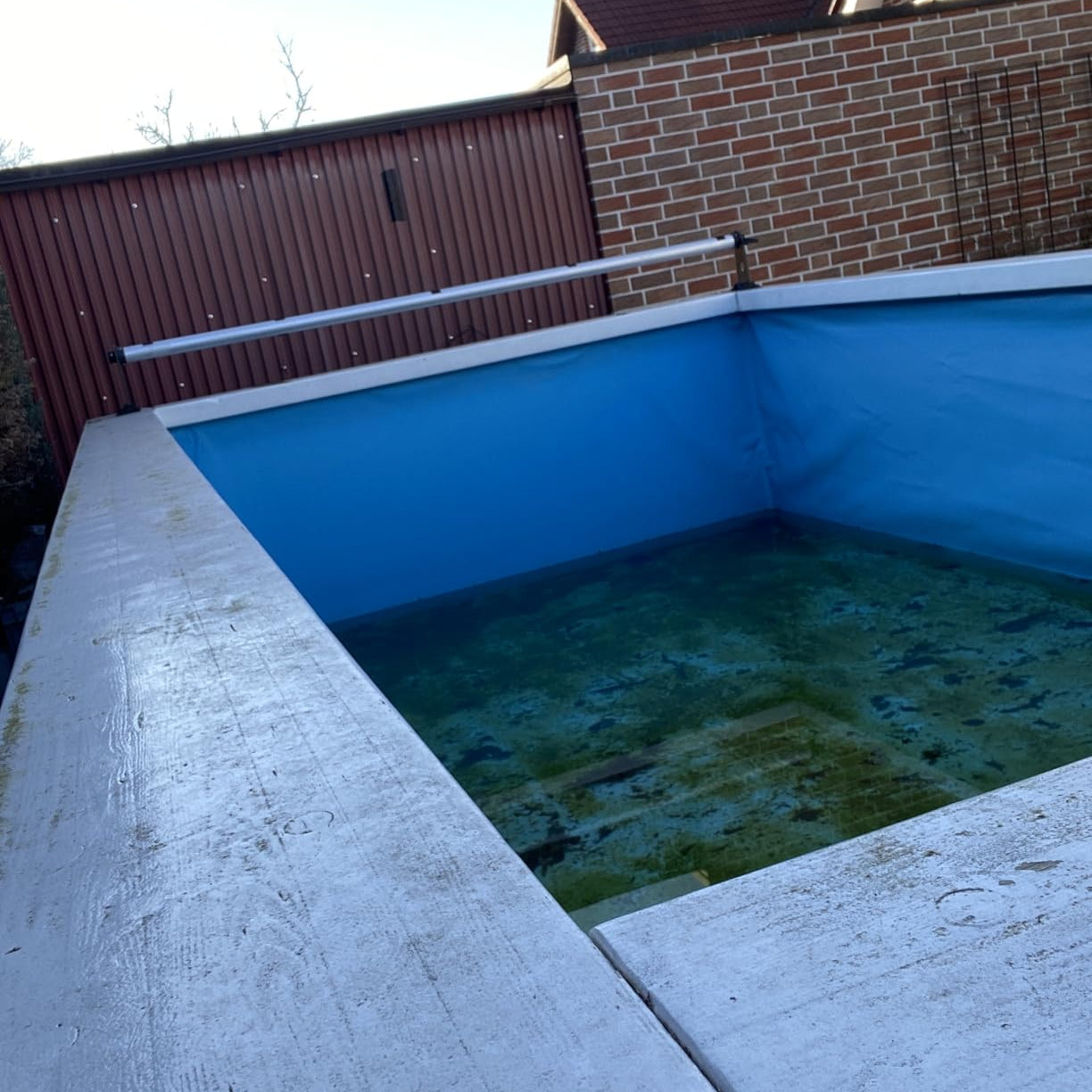 Aussenpool mit blauem Becken, teilweise gefuellt mit Wasser, umgeben von Betonrand und Ziegelwand im Hintergrund