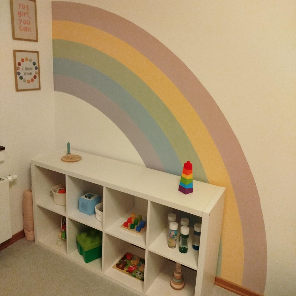 Wandgestaltung im Kinderzimmer mit großem Regenbogen in Pastellfarben über weißem Regal mit Spielzeug.