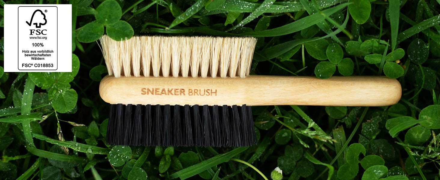 Sneaker Brush 2-in-1 Reinigungsbürste mit FSC-zertifiziertem Holzgriff und hellen Naturborsten sowie schwarzen Polyamidborsten auf grünem Kleeuntergrund als Symbol für Nachhaltigkeit.