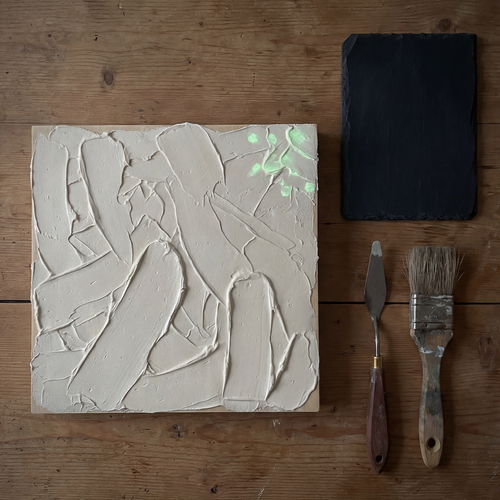 Quadratische Leinwand mit aufgetragener Strukturpaste in groben Spachtelstrichen, daneben Spachtel, Pinsel und eine dunkle Musterplatte auf einem Holztisch.