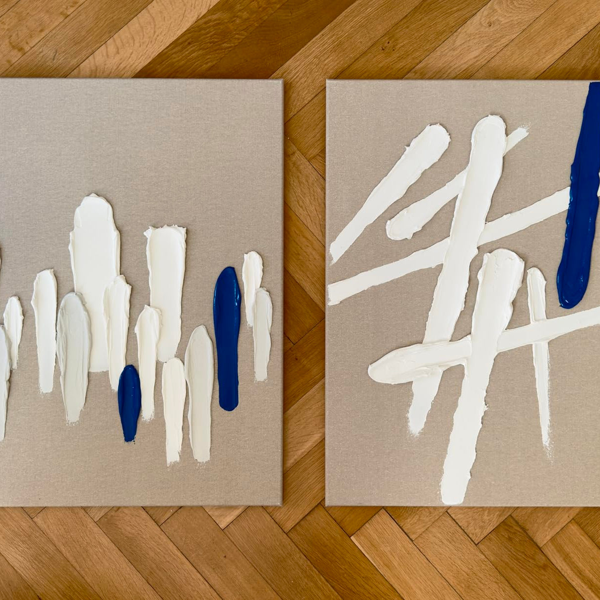 Zwei Leinwände mit abstrakten Strukturen aus weißer Strukturpaste und blauen Akzenten auf Holzfußboden.